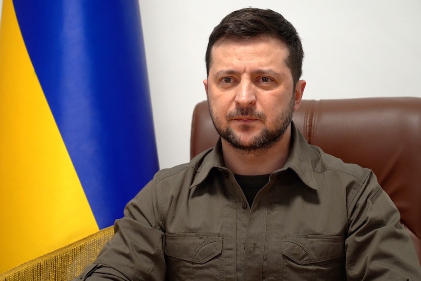 Volodymyr Zelensky sits in a brown leather chair with a Ukrainian flag behind him