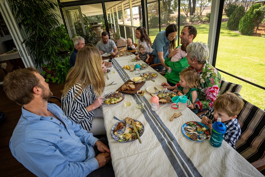 A family gathers around a long table for lunch. Food sits on top of a white table cloth as the family laugh and chat. 