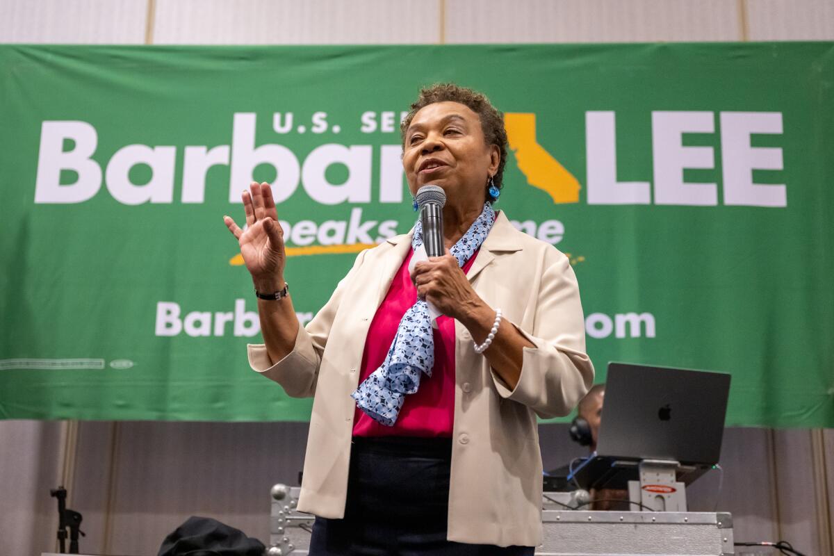 Rep. Barbara Lee gesturing as she speaks into a microphone in front of a large green campaign banner with her name in white