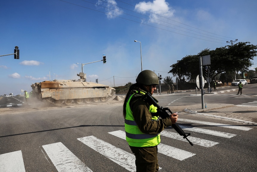 A soldier carrying a gun and wearing a helmet and hi vis vest crosses a road with a tank in the background