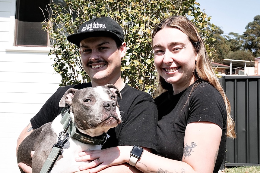 Man and woman smile at the camera while hugging a dog. 