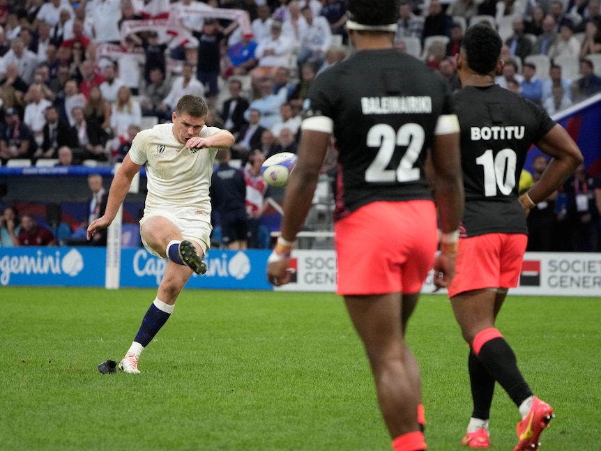 An English rugby union player sends a kick towards the posts as a line of Fijian players watch.