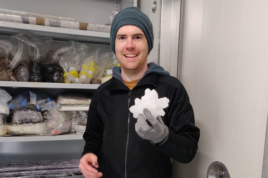 A man wearing a jacket and beannie smiles as he stands in front of a freezer and holds a large hailstone.