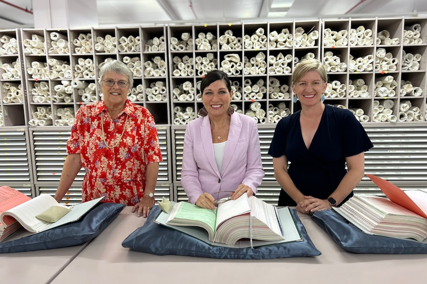 Ms Enoch, Dr Kerr, and Ms Howard smiling while standing in front with old archive books.
