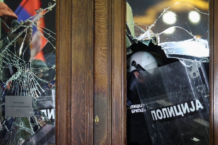 A smashed window and through it you can see a riot policeman with a shield 