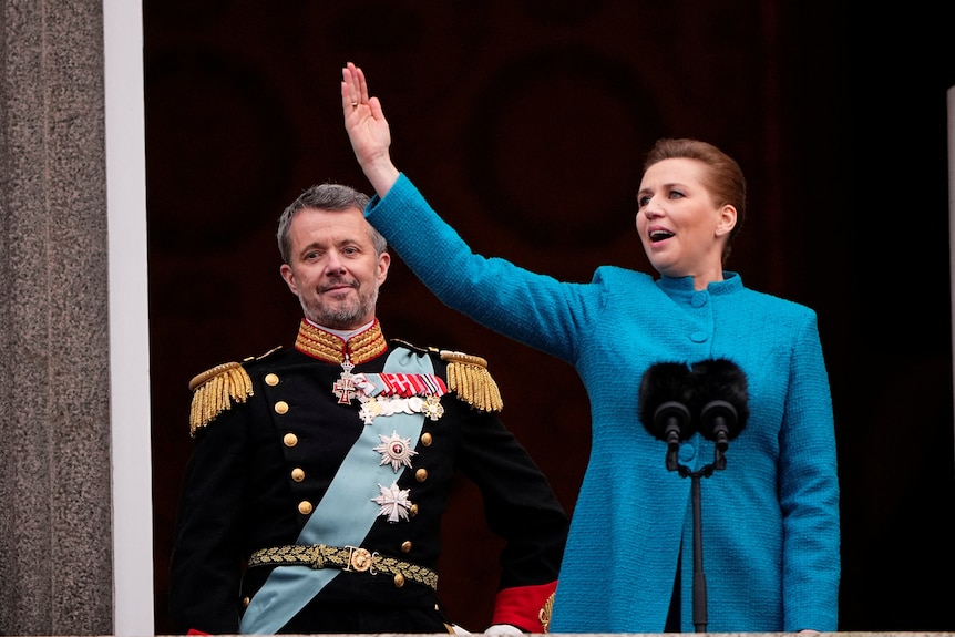 Mette, dressed in a blue suit, thrusts her right arm into the air as she speaks with Frederik standing behind her