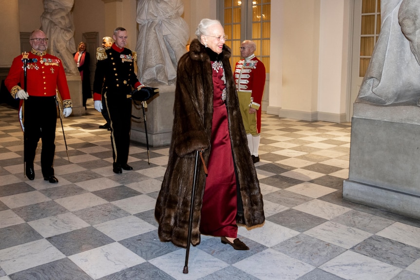 Queen Margrethe walks through a room wearing a full length fur coat and red gown.