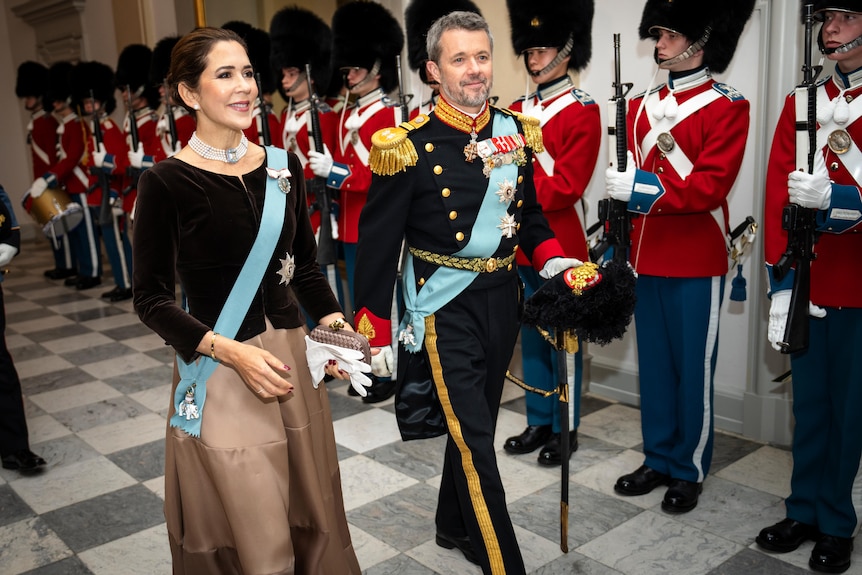 Princess Mary and Prince Frederik walk through a room side by side with guards in the background.