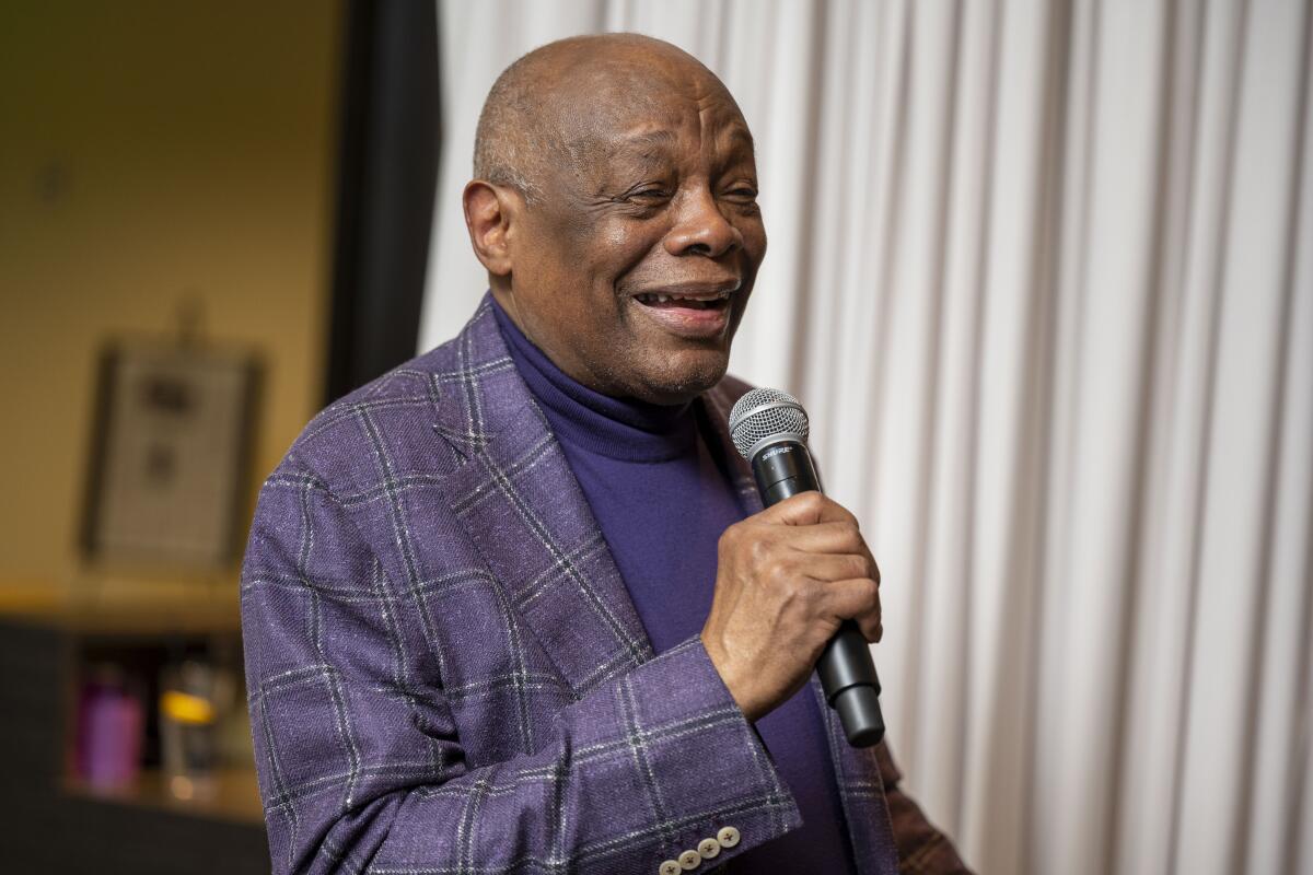 Former San Francisco Mayor Willie Brown in a purple turtleneck and checked purple jacket holds a microphone