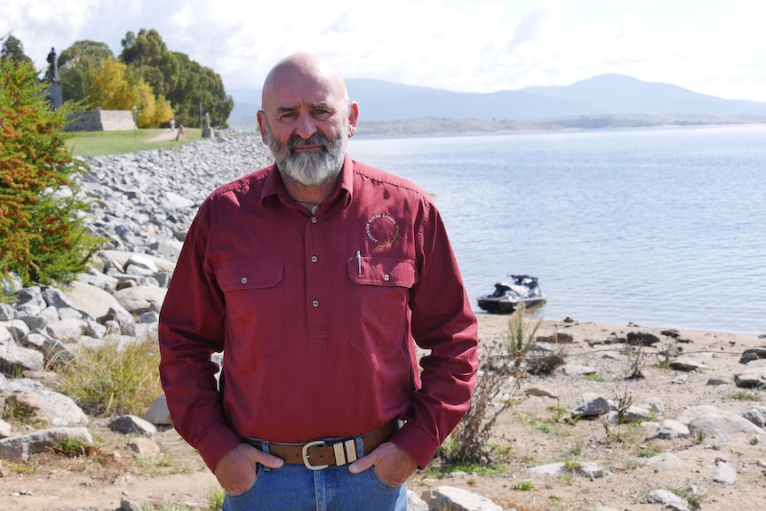 A man in a red button up shirt and blue jeans stands with hands in his pocket by a lakeside