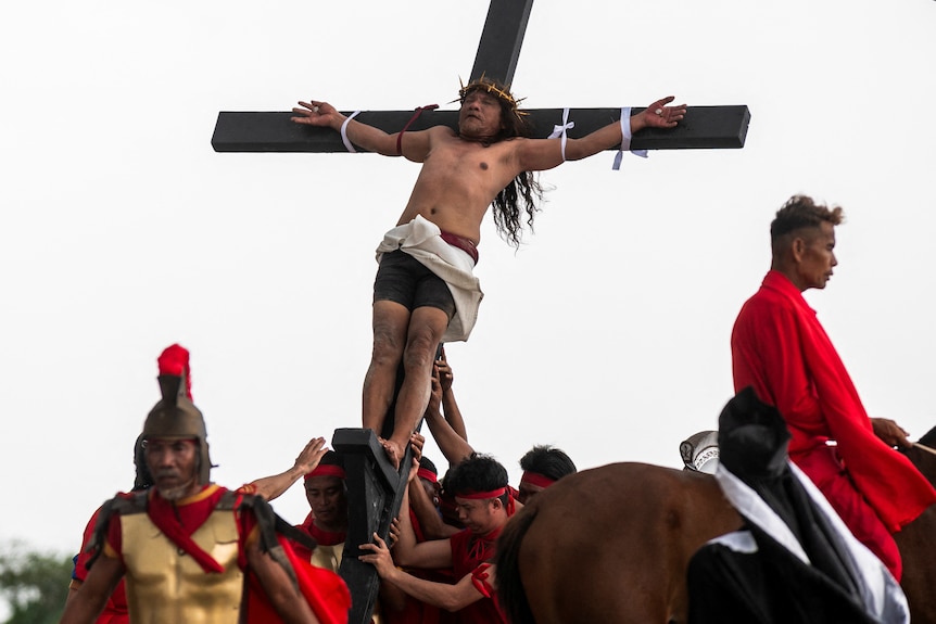 Men push a cross up. On the cross, a man wearing a white robe around his waist is nailed by his hands.