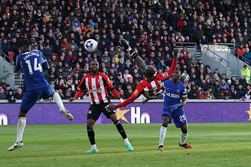 A Premier League footballer twists in mid-air after volleying the ball over his head towards the goal as players watch.