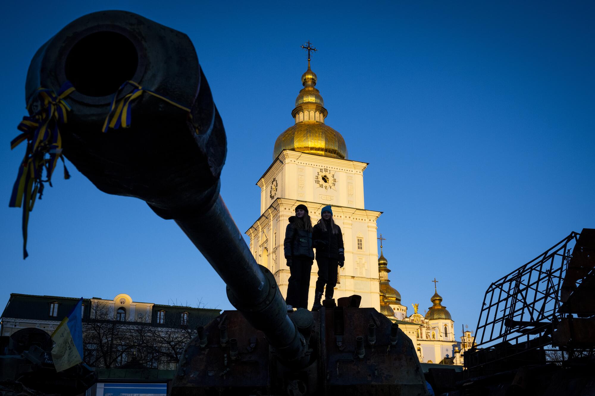 People stand in shadow near a tank displayed in Kyiv. A gold-domed monastery is lighted up in the background