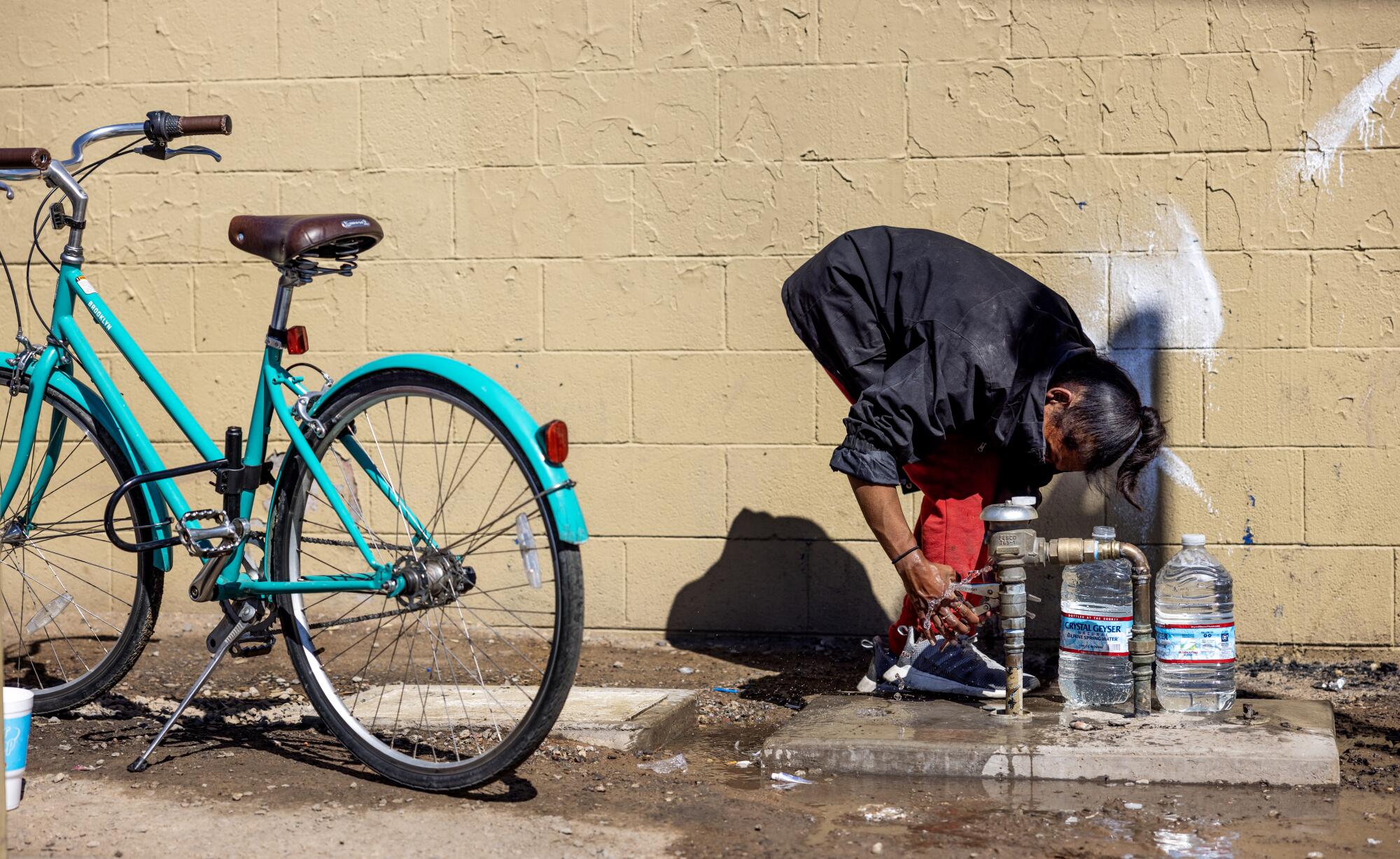 A person with plastic water jugs bends over near a parked bicycle to open a valve against a wall