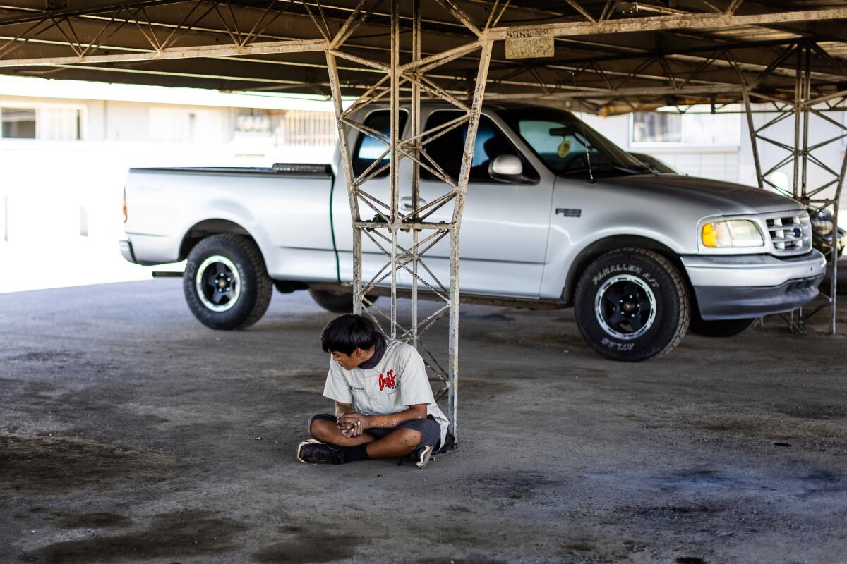 A young man sits cross-legged in a covered parking area near a pickup, turning his head away