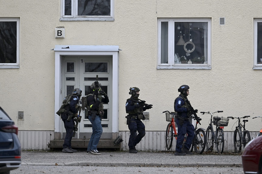 Four police officers wearing special gear and carrying rifles stand on the side of a building.
