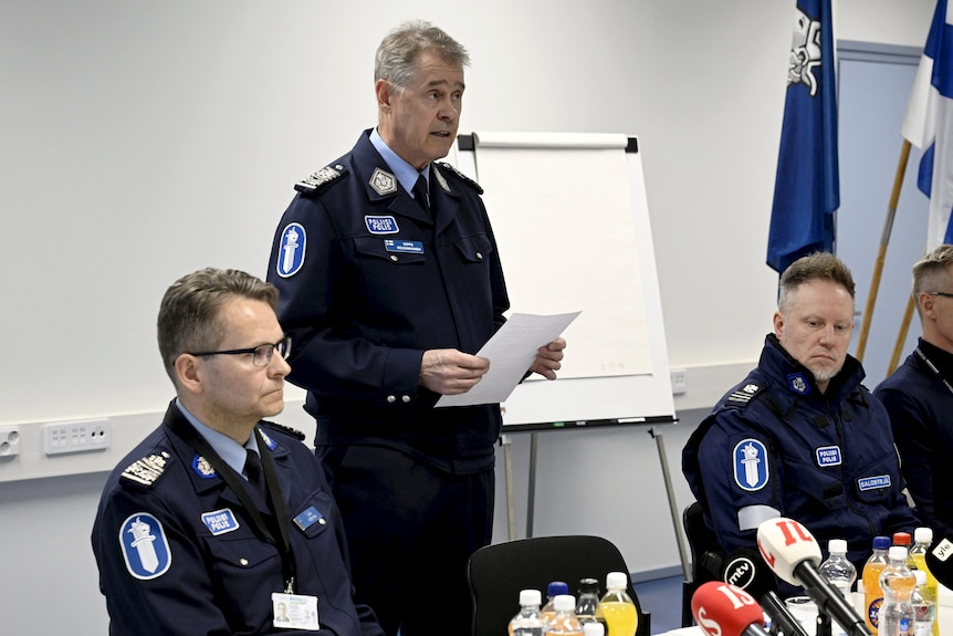 Police men wearing uniforms behind a table at a press briefing with one standing up and holding a paper