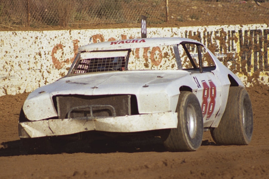 A white sedan with large tyres and 88 in big red letters on the side, on a red dirt speedway.