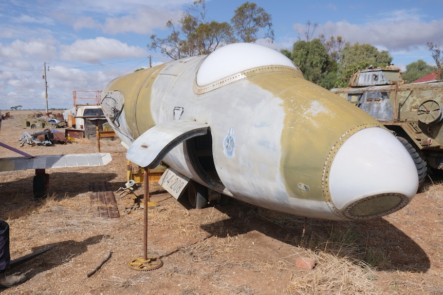 The nose of a grey and green camouflage plane with no wings sits on dry grass with a tank behind 
