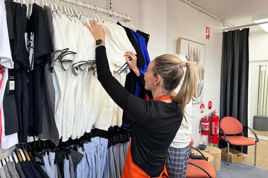 A woman in a clothing shop sorts through garments hanging from a wall.