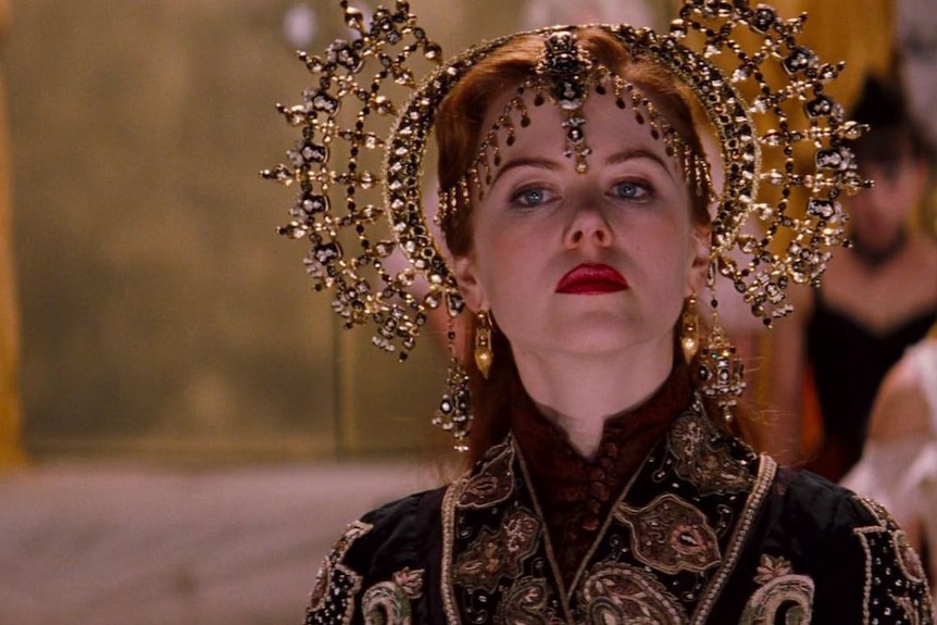 Nicole Kidman wears an intricate shining gold headpiece and looks to the right while walking through a room.