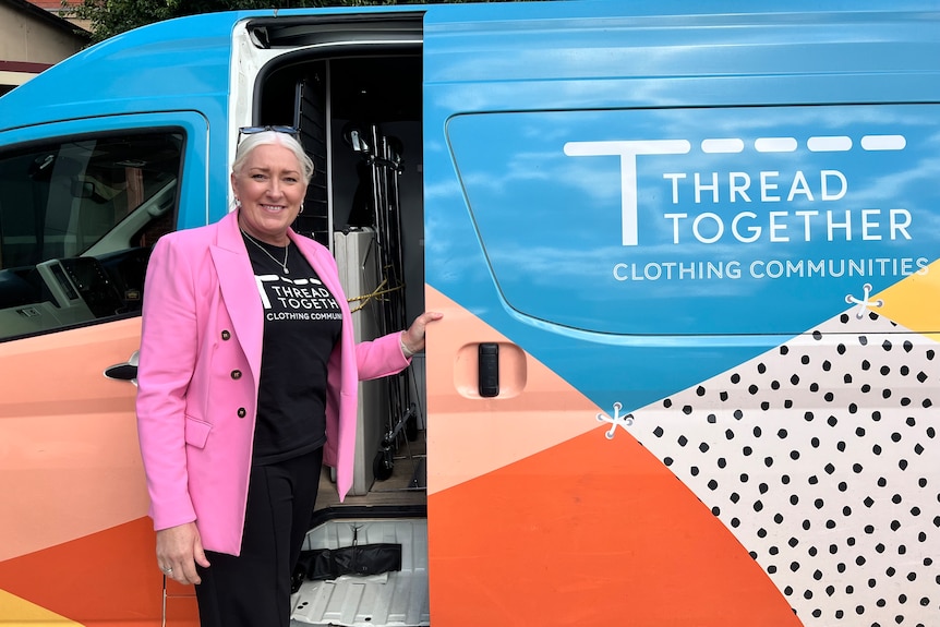 A smiling woman in a brightly-coloured blazer stands in front of a van.