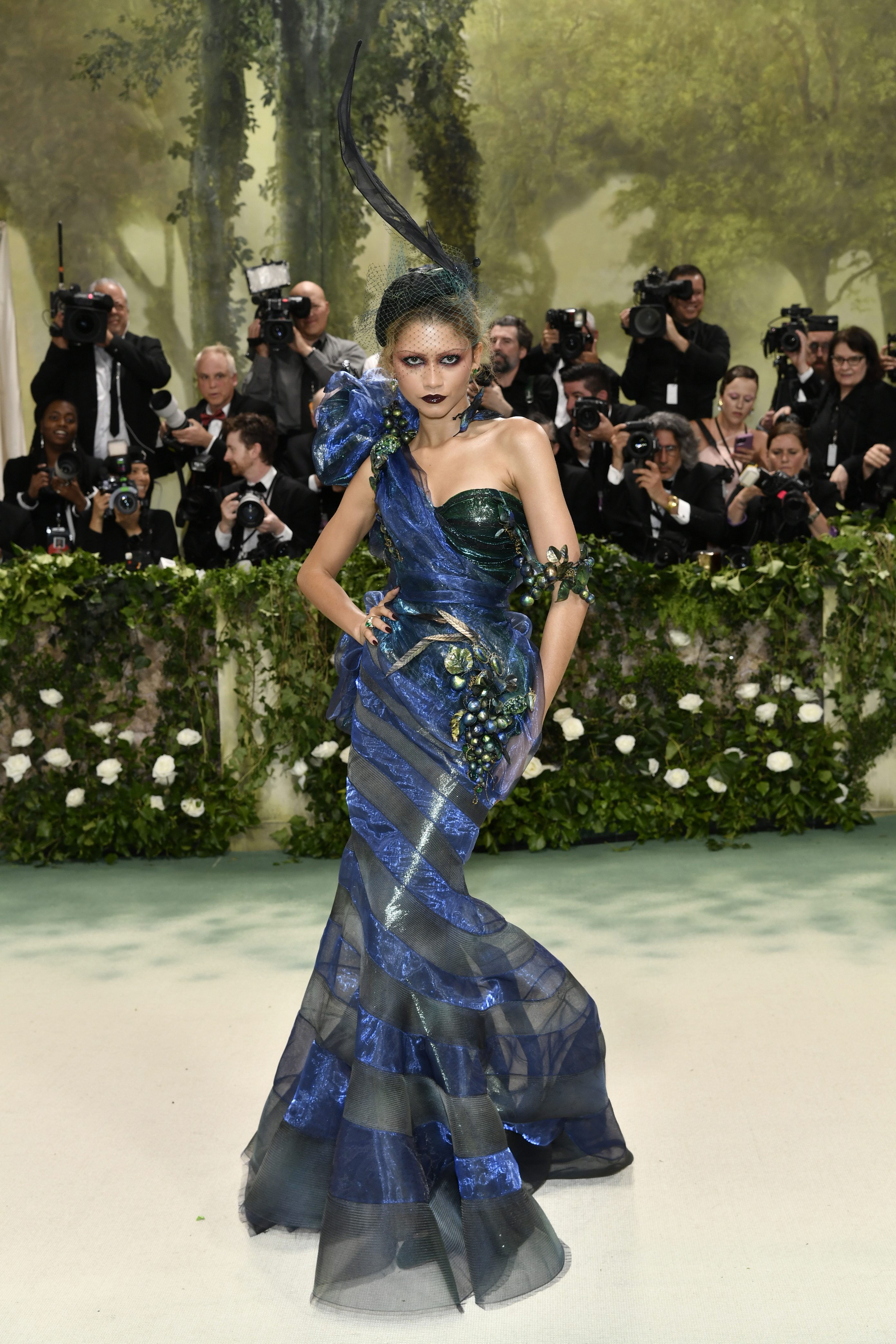 Zendaya wearing a dark green and blue fitted mermaid dress with a dark green headpiece with a tall feather-like detail 
