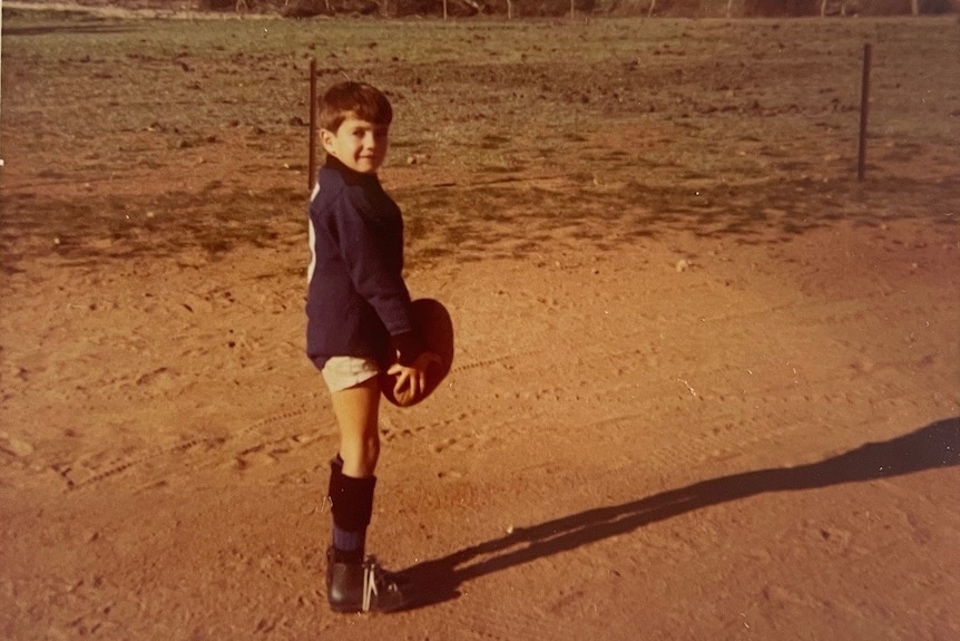 Small boy wears jumper, shorts and boots holding a football