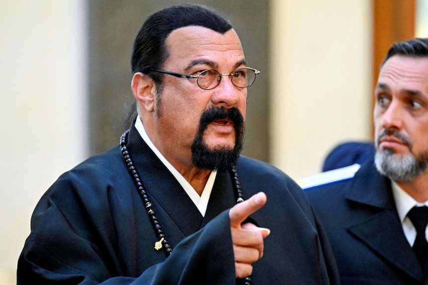 Actor Steven Segal wears a black shirt with a beaded necklace and glasses, pointing at someone out of shot