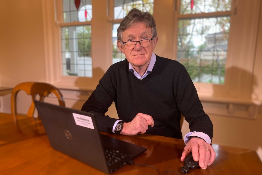 Alan Oster, in glasses, collared shirt and jumper, sits at a wooden dining table with his laptop open in front of him