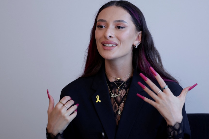 A close up of Eden Golan speaking. She's got long pink fingernails and is wearing a pin with a yellow ribbon on her black blazer