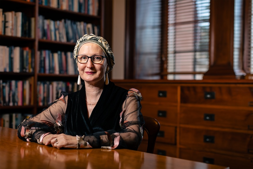 Angela O'Keeffe, a woman in her 60s wearing a headscarf and glasses, sits behind a desk, in front of a bookshelf.