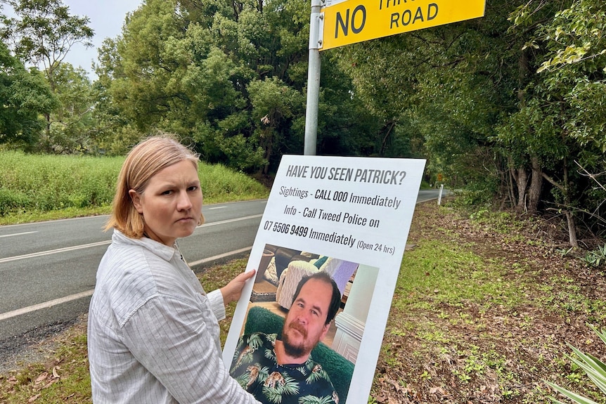 A woman holds a large poster with a picture of a man on the side of a regional road. 