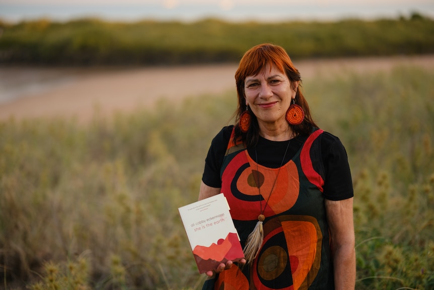 Ali Cobby Eckermann, a 60-year old Aboriginal woman, stands near a beach, holding her book She Is the Earth