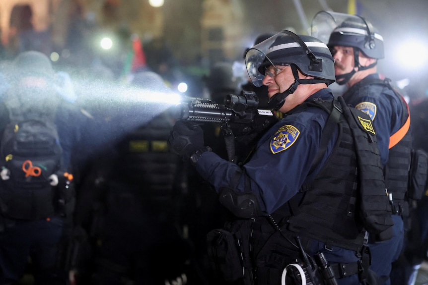 A policeman points a weapon toward something out of shot while standing in front of a barricade