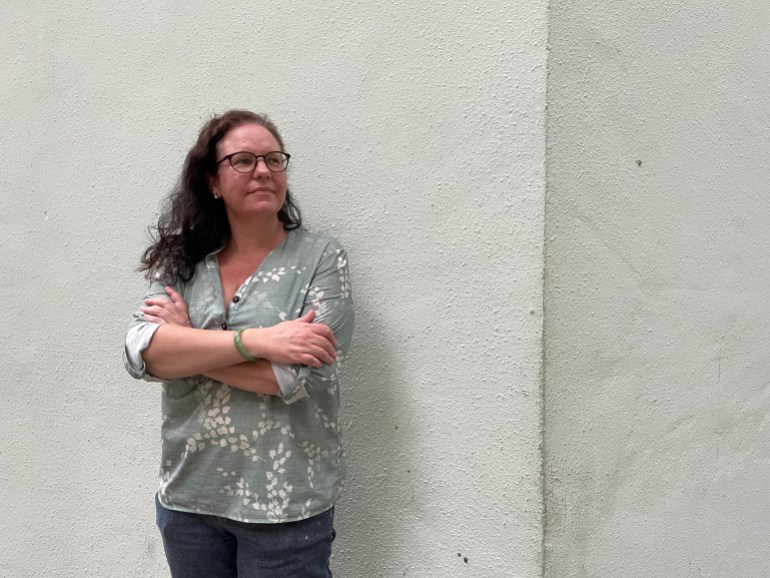Chloe Martin. She has curly medium length hair and is wearing glasses. She is standing against a white wall