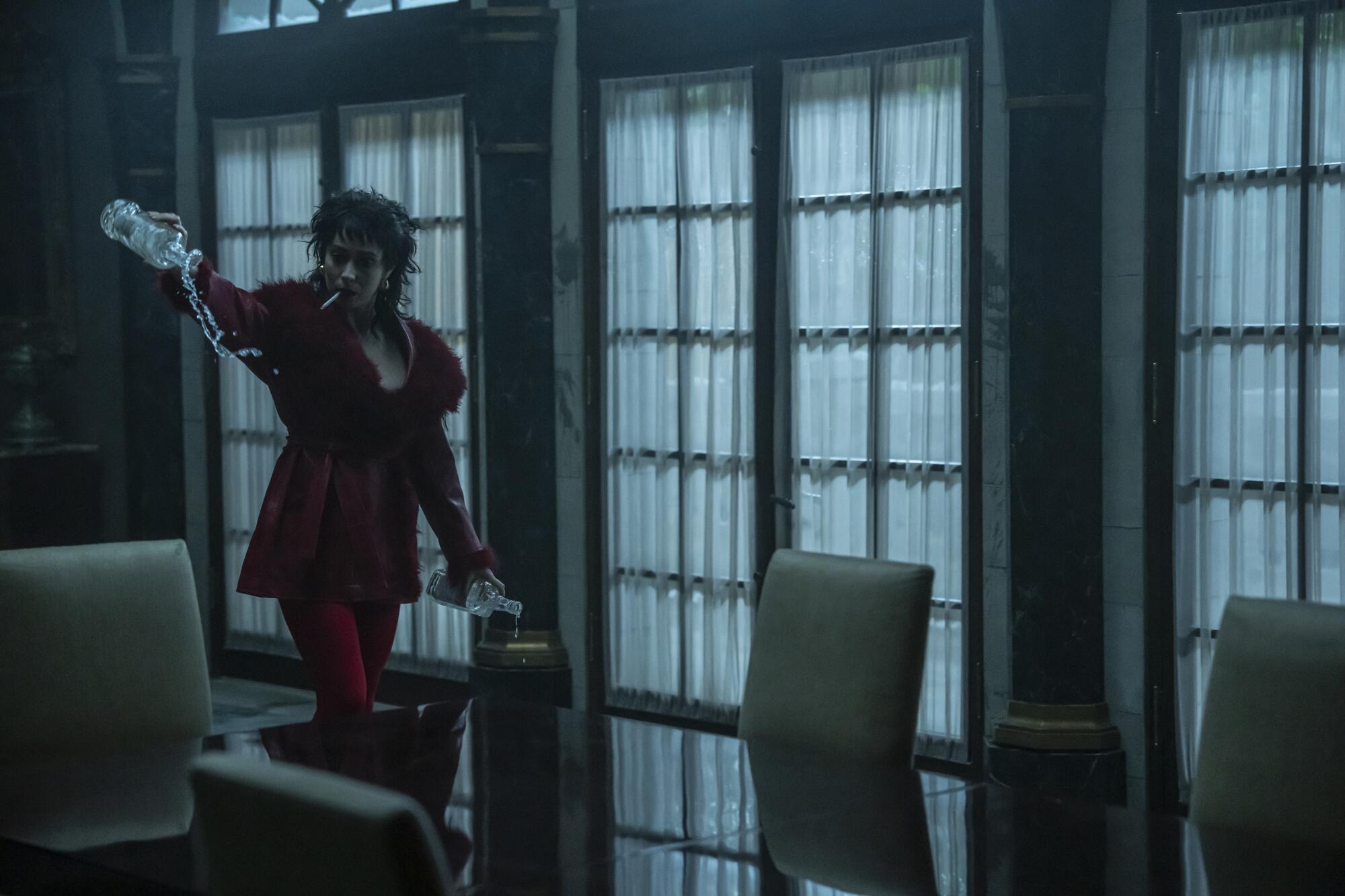A woman in a red coat smoking a cigarette pours a bottle of liquor on the floor.