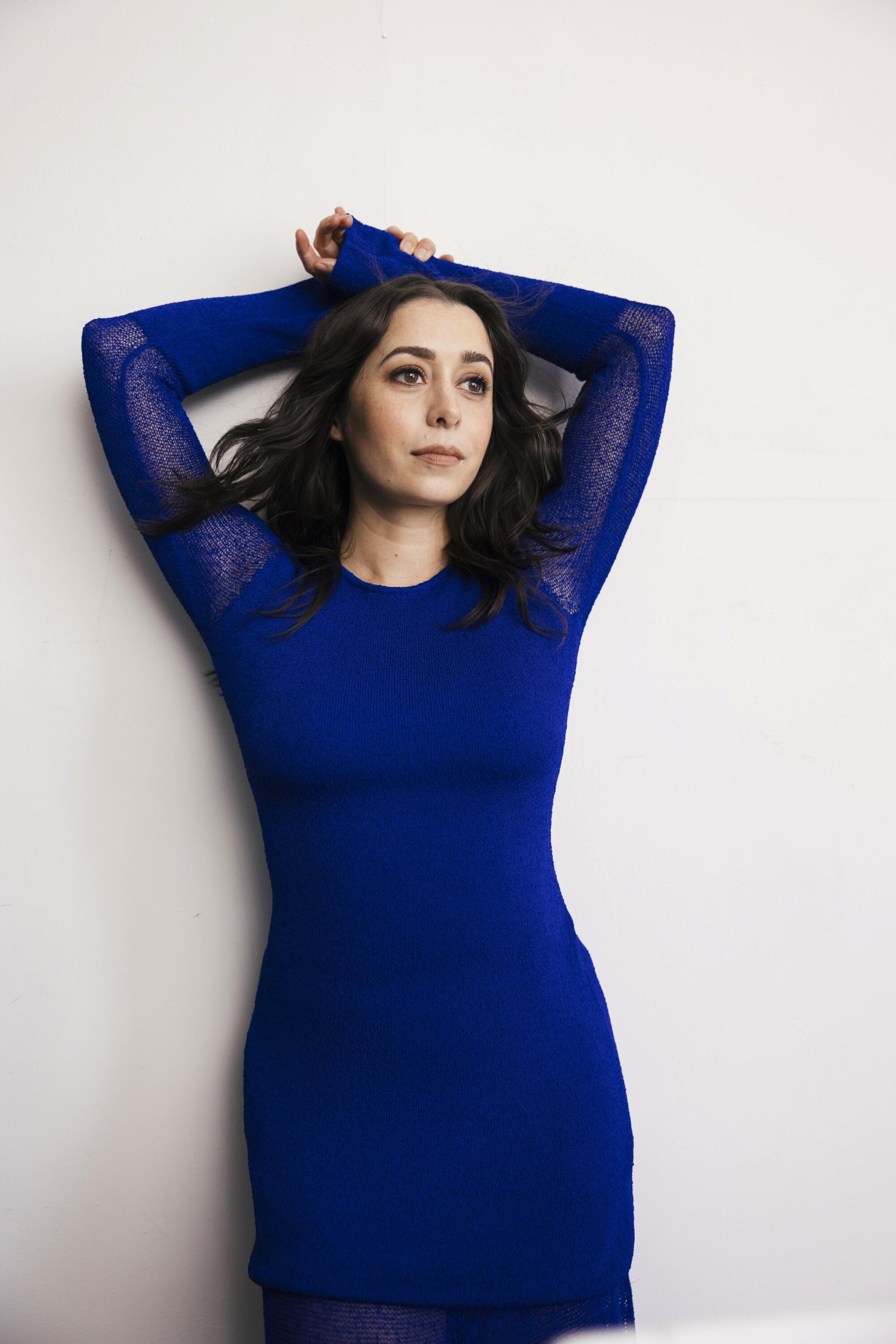 A woman in a blue long-sleeve dress poses against a wall with her hands crossed above her head.
