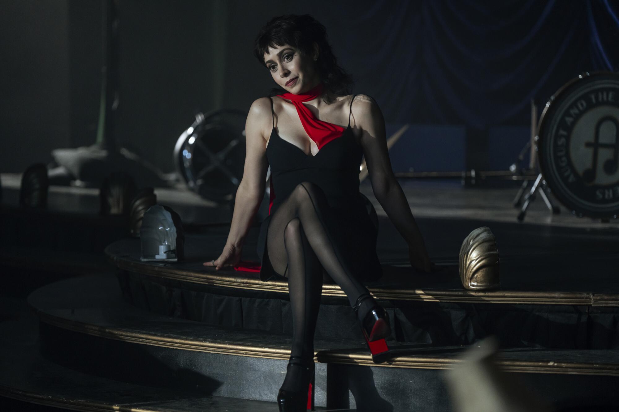 A woman in a black cocktail dress and red scarf sits and leans on a set of black stairs.