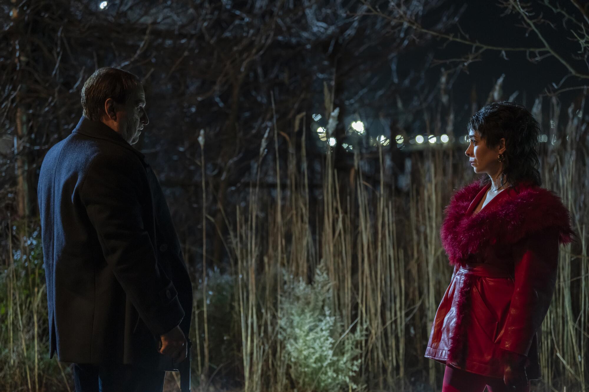 A man in a black suit stands in a field holding a gun by his side and facing at a woman in red coat.