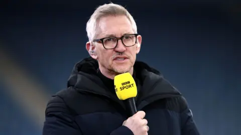 PA Gary Lineker, wearing a black coat, holds a BBC Sport microphone and wears headphones as he presents football coverage on TV