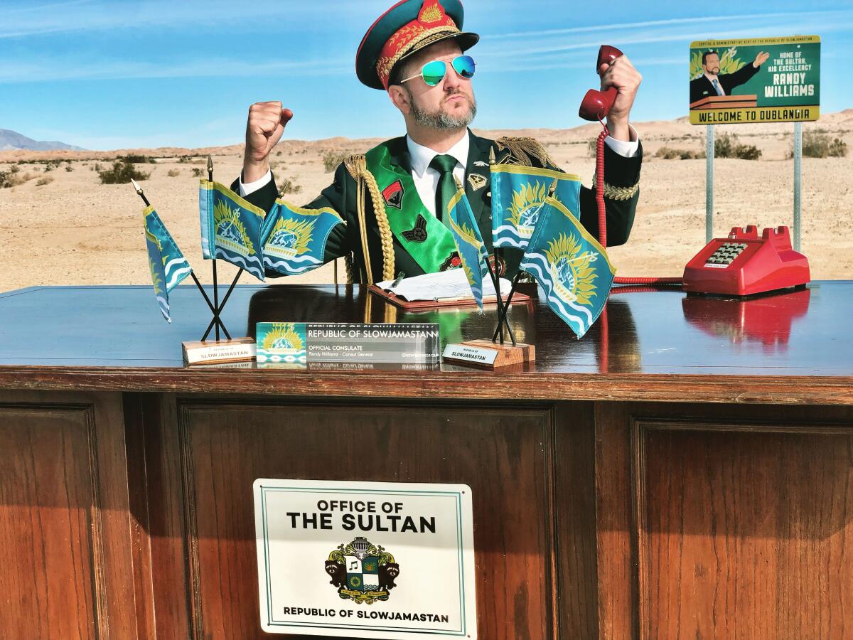 A man in a military uniform clenches his fists as he sits at a desk with small flags on it with the desert in background.