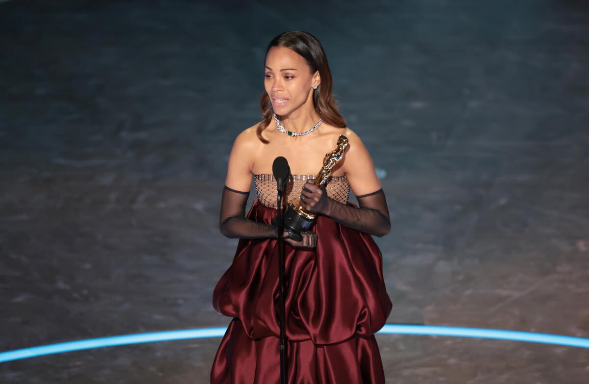 A woman in maroon dress and black mesh gloves on a stage speaking at a mic and holding a statuette.