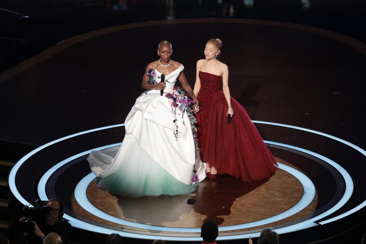 A woman in white dress adorned with flowers holds the hand of a woman in a red sparkling dress on a stage.
