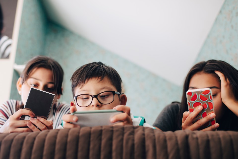 Three siblings lying down on a couch using digital devices.
