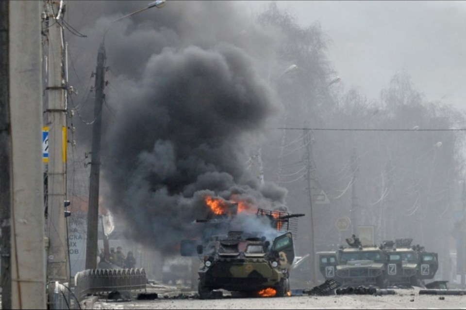 Burning military vehicles amidst smoke and debris on a city street.