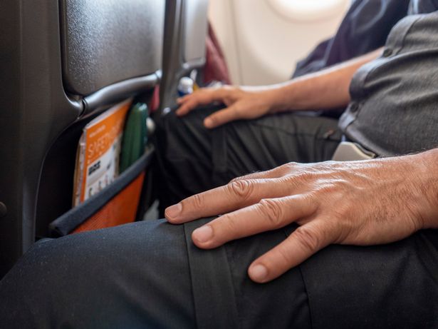 A man resting his hands on his lap as he sits aboard a commercial airline flight. The seat safety buckle is secured across his lap, his knees almost touching the seat in front and his legs apart to make more space for himself, as he sits in the middle seat of a row of seats. He is wearing a grey short sleeved shirt and black trousers. Light is coming in from the nearby cabin window. The aeroplane safety manual and a green glasses case are stored in the pocket of the seat in front of him.