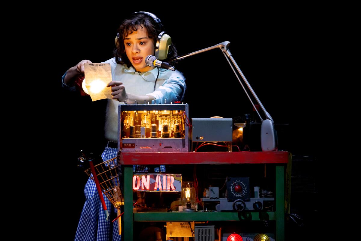 A young woman in headphones at a radio broadcasting setup holds a flashlight on a piece of paper