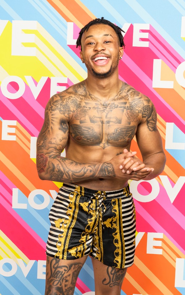 A smiling man with tattoos, wearing patterned shorts, poses in front of a "Love Island" backdrop.