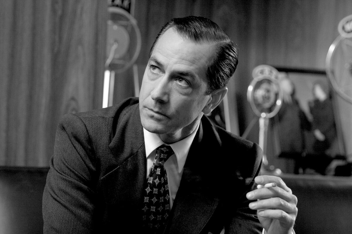 A black and white photograph of a man wearing a suit and tie and holding a cigarette